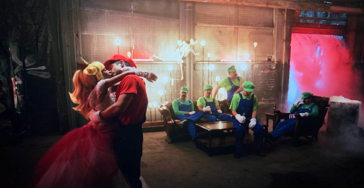 Cinque uomini vestiti da luigi rimangono tristi e in disparte, mentre un ragazzo vestito da Mario bacia una ragazza con gli abiti della principessa Peach.