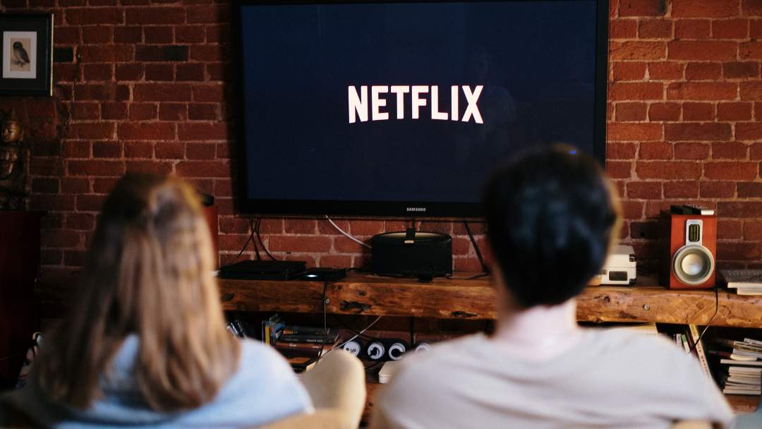 Um casal sentado de costas em um sofá em frente à TV com a tela inicial do Netflix