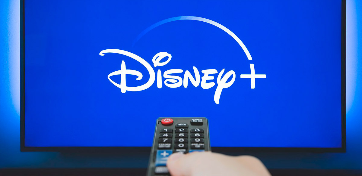Una mano punta il telecomando sul televisore dove c'è il logo Disney+