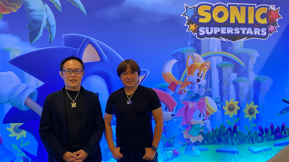 Takashi Iizuka e Naoto 'Big Island' Ōshima in posa davanti a una grafica di Sonic Superstars