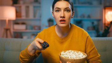 donna che guarda la tv con il telecomando ei popcorn indossa un maglione giallo e ha la faccia sorpresa