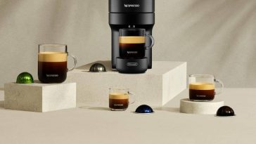 macchina nespresso sul piedistallo con tazzine di caffè e capsule su sfondo monocromatico