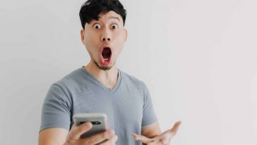 uomo sorpreso che guarda in camera con in mano uno smartphone grigio