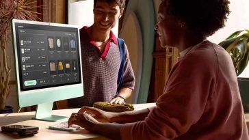 Persona seduta alla scrivania che digita su tastiera di un iMac con un'altra persona dietro la scrivania in piedi che sorride