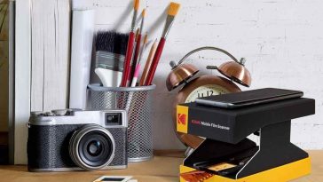 scanner portatile Kodak appoggiato sulla scrivania accanto a una macchina fotografica e davanti a un bicchiere con pennelli, una sveglia e un libro