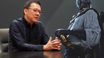 Hidetaka Miyazaki seduto a una tavolo che parla con le braccia appoggiate sul tavolo e di fianco la figura del protagonista di The Duskbloods, un uomo con camicia bianca, pantaloni neri, caschetto bianco e pistola nella fondina