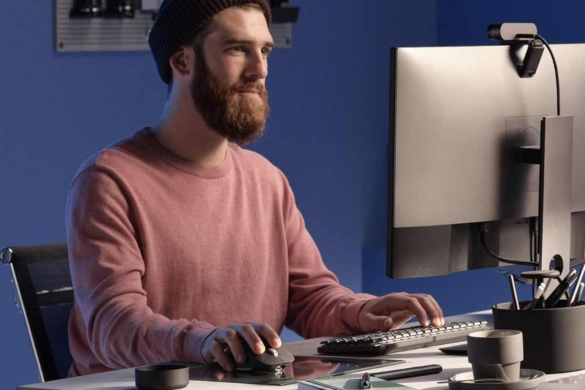 Ragazzo con barba, berrettino nero, maglia salmone, davanti a schermo del computer con mouse in mano