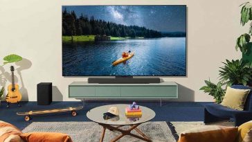 televisore LG OLED appeso alla parete in un salotto con soundbar integrata, di fronte a un divano giallo e in mezzo c'è un tavolino di vetro con alcuni oggetti sopra e uno skateboard sul pavimento