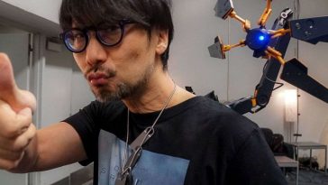 hideo kojima che fa faccia buffa con alle spalle congegno elettronico attaccato con braccio elettronico