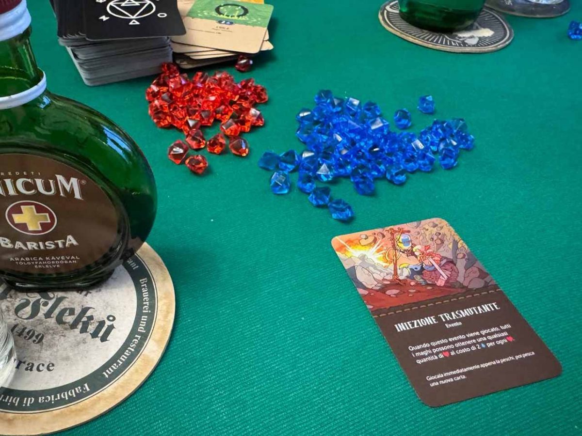 Dettaglio del centro-tavolo di Not Enough Mana con una carta Evento scoperta, segnalini, mazzo da pesca e pila degli scarti (con alcol di contorno)