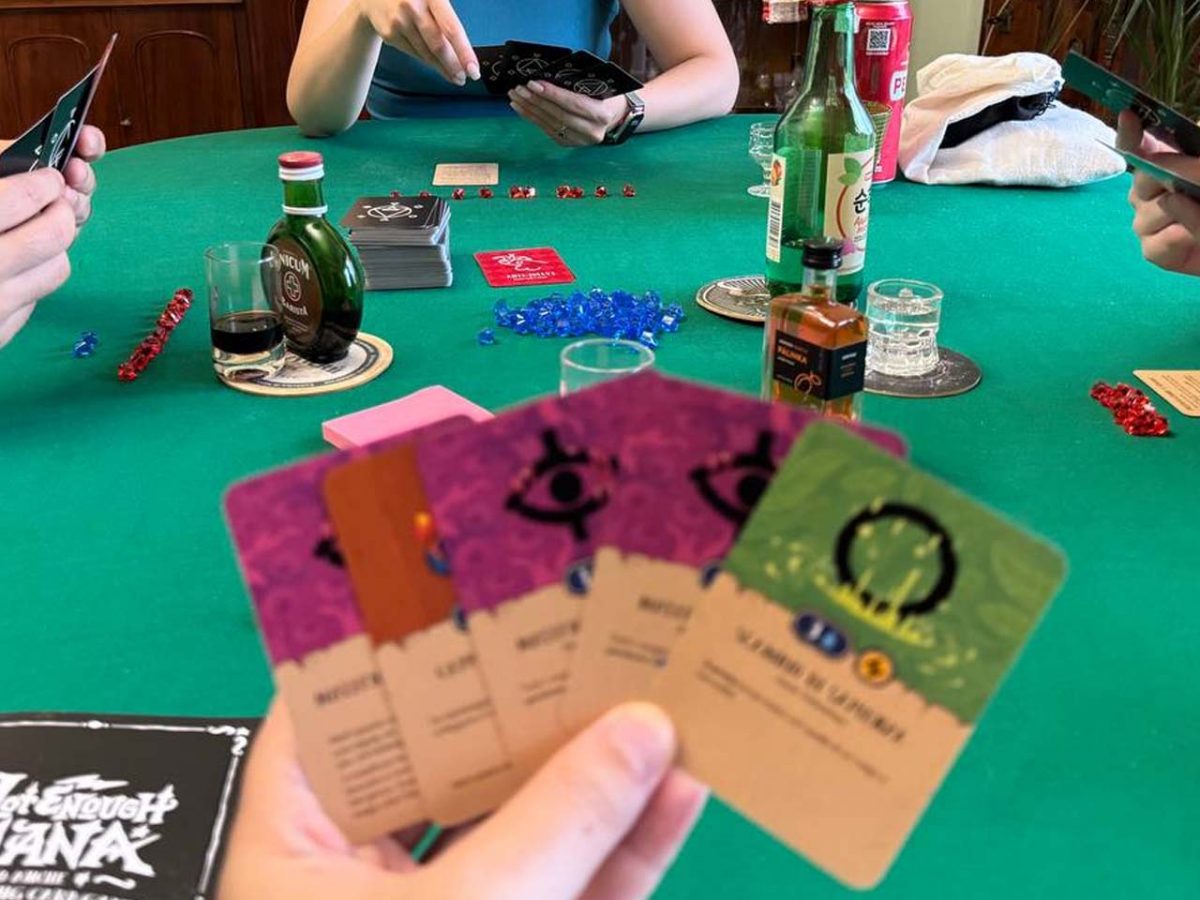 Mano del giocatore con carte di Not Enough Mana offuscata, focus sul tavolo da gioco dietro imbandito con alcol e segnalini