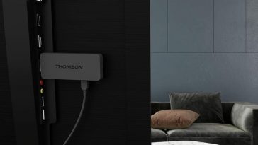 THOMSON Google TV Streaming Stick 140 collegata sul retro di televisore, a destra salotto con tavolo, divano e cuscini