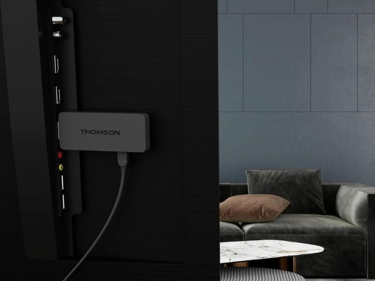 THOMSON Google TV Streaming Stick 140 collegata sul retro di televisore, a destra salotto con tavolo, divano e cuscini