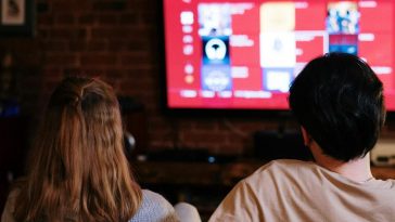 Una coppia di spalle sul divano che sceglie un contenuto su una piattaforma di streaming sulla tv