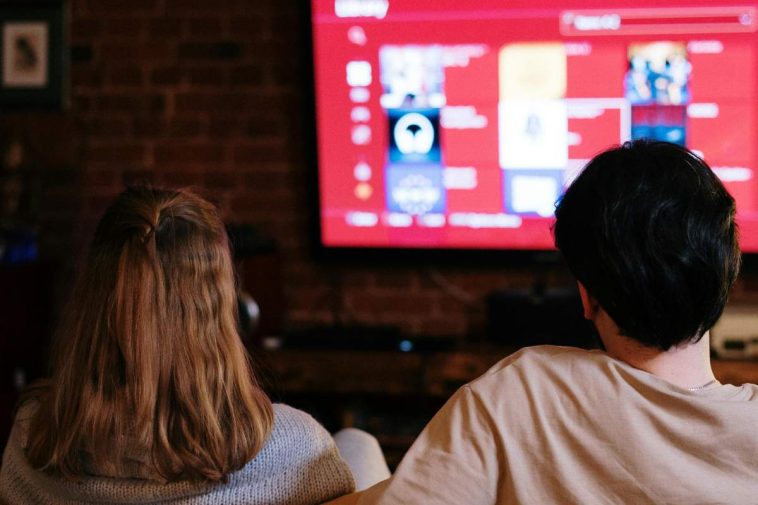 Una coppia di spalle sul divano che sceglie un contenuto su una piattaforma di streaming sulla tv