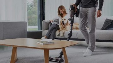 Un uomo passa l'aspirapolvere mentre la moglie e il cane stanno sul divano