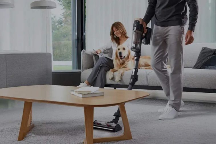 Un uomo passa l'aspirapolvere mentre la moglie e il cane stanno sul divano