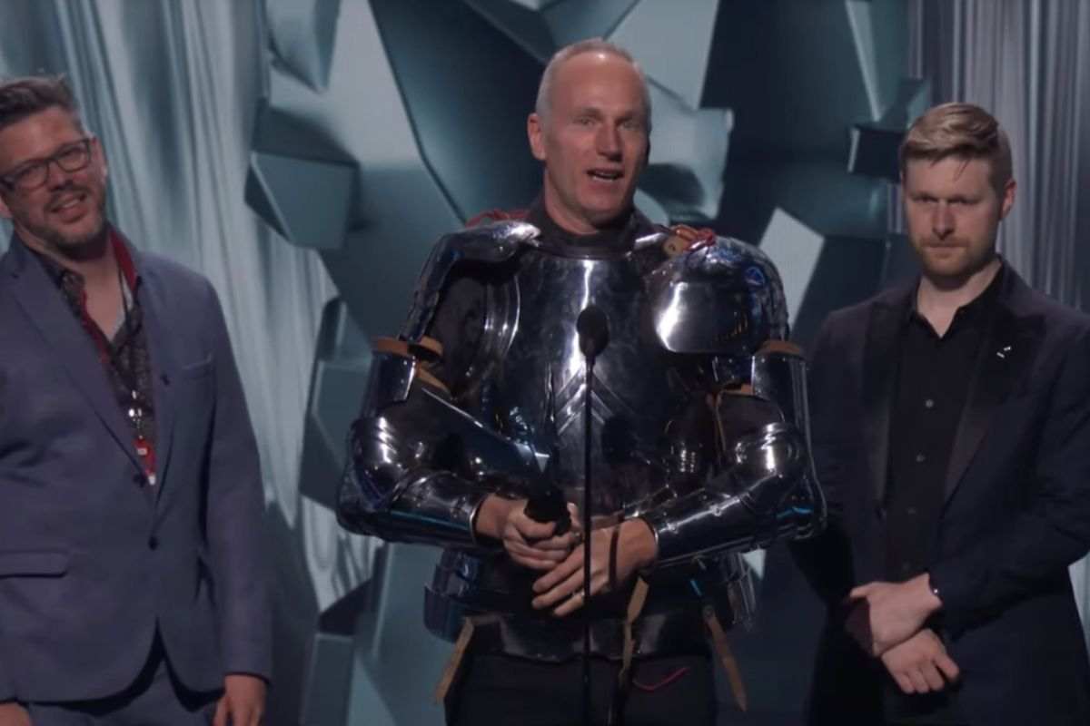 uomo con capelli banchi in armatura di ferro medievale con mantello rosso con n mano statuetta premio, accanto due uomini in abiti civili