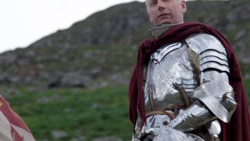 uomo con capelli banchi in armatura di ferro medievale con mantello rosso, detro montagna