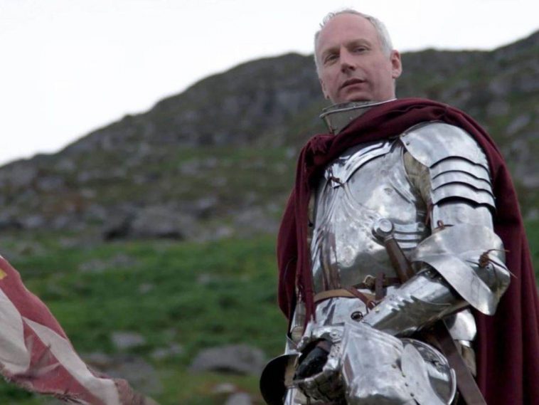 uomo con capelli banchi in armatura di ferro medievale con mantello rosso, detro montagna
