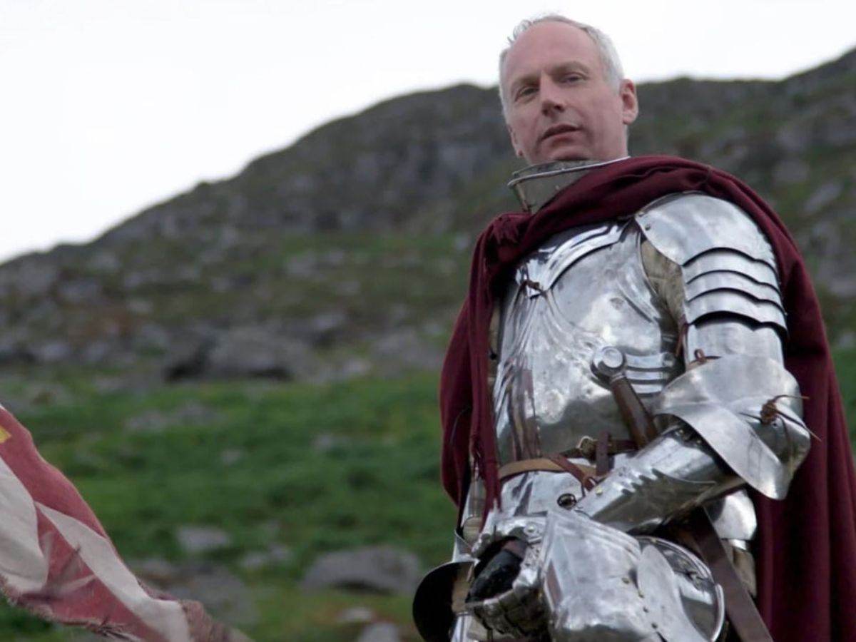 uomo con capelli banchi in armatura di ferro medievale con mantello rosso, detro montagna