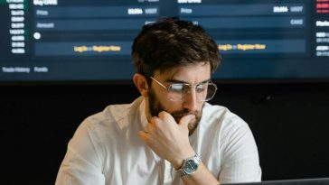 Un ragazzo con gli occhiali al computer con alle spalle delle proiezioni finanziarie