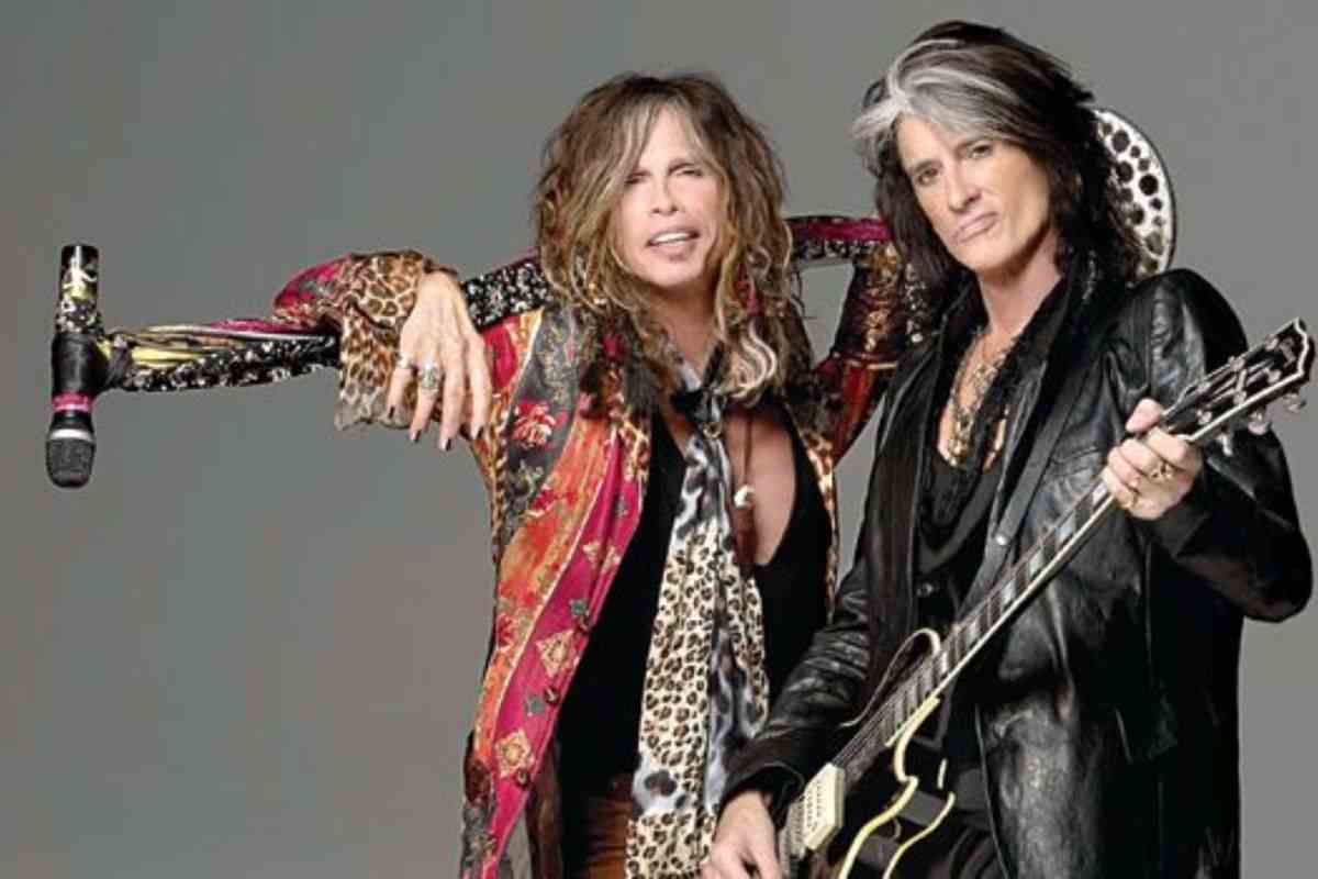 steven tyler con casacca rossa e microfono su asta attorno al collo, joe perry con cappotto grigio e chitarra nera in spalla