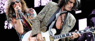 chitarrista ( joe perry) con capelli neri e bianchi, giacca maculata e chitarra bianca con immagine di ragazza, cantante (Steven Tyler) con occhiali da sole micrfono in mano