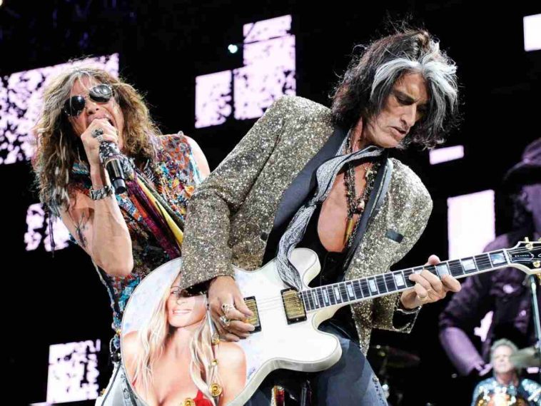 chitarrista ( joe perry) con capelli neri e bianchi, giacca maculata e chitarra bianca con immagine di ragazza, cantante (Steven Tyler) con occhiali da sole micrfono in mano