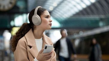 ragazza con capelli ricci, cellulare, cappotto beige, cuffie bianche alla stazione dei treni