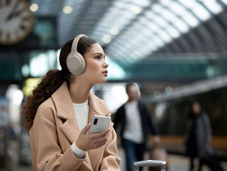 ragazza con capelli ricci, cellulare, cappotto beige, cuffie bianche alla stazione dei treni