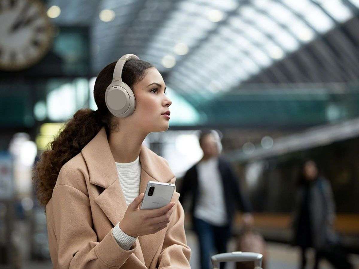 ragazza con capelli ricci, cellulare, cappotto beige, cuffie bianche alla stazione dei treni
